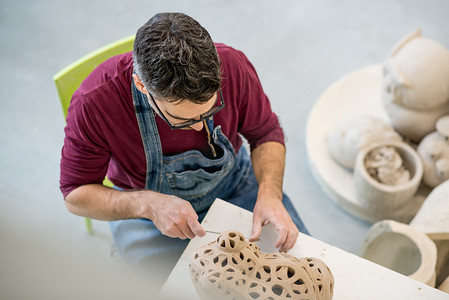 陶艺古文化摄影照片_明亮陶瓷工作室中穿着围裙的陶艺家用生粘土雕刻雕像的俯视图。
