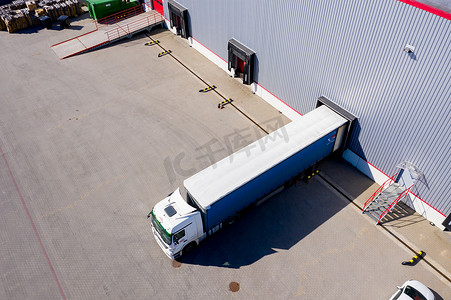 空中拍摄的带有半挂车的卡车离开工业仓库/仓储大楼/装货区，许多卡车正在装卸货物