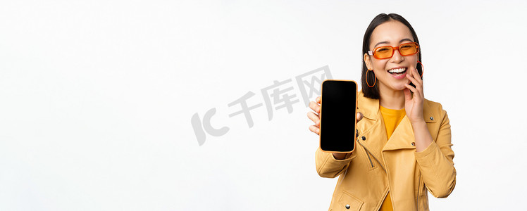 热情的亚洲女模特，在手机屏幕上展示智能手机应用程序界面、网上商店或网站，站在白色背景上