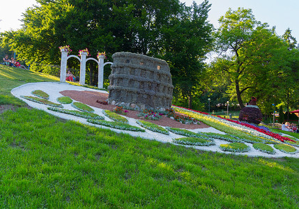 公园里的游客坐在斜坡上，凝视着花坛中央的一座堡垒形状的雕塑