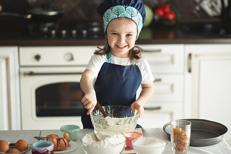 一个穿着围裙、戴着厨师帽的可爱小女孩正在用木铲搅拌面团，一边做饭一边看着镜头微笑