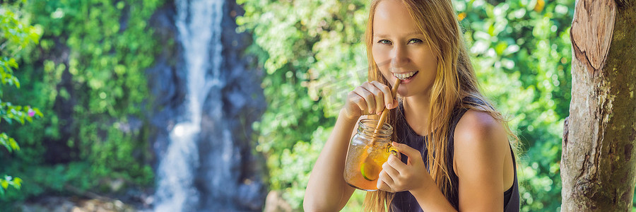 饮料banner摄影照片_一个美丽的女人喝冰茶的特写肖像图像，在绿色自然和瀑布花园背景中感到快乐 BANNER，长格式