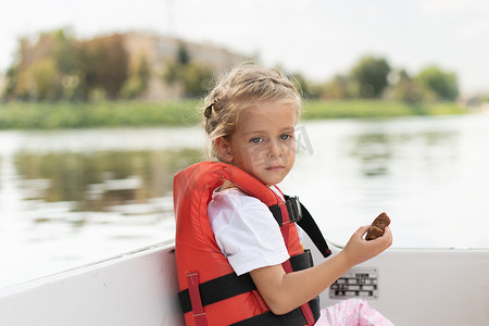 游船河摄影照片_小女孩穿着救生衣在城市河上乘坐游船阳光明媚的夏日白人女孩在船上游泳