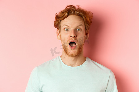 炫酷红色摄影照片_惊讶的红发男人对酷炫的促销优惠做出反应，扬起眉毛，喘着粗气，敬畏地盯着镜头，站在粉红色背景上