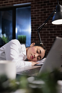 办公室休息区摄影照片_加班后睡在办公室工作区桌子上的疲惫女商人的肖像照片。