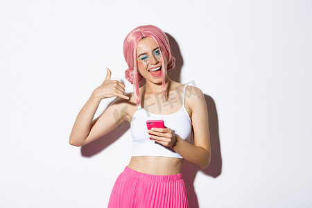 欢快的派对女孩打着电话信号，对着镜头眨眼调情，拿着智能手机，站在白色背景的粉色假发中