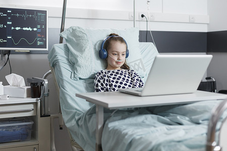 看病床摄影照片_年轻耐心的女孩坐在儿科医院的病床上看有趣的卡通片。