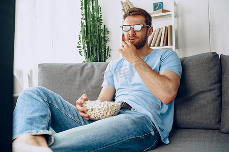 戴着 3D 眼镜的大胡子男人坐在沙发上，用爆米花看电影
