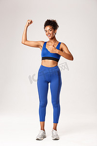 全长身穿蓝色运动服的强壮迷人的非洲裔美国女运动员，竖起大拇指和弯曲的二头肌，推荐健身房锻炼，良好的训练课程，白色背景