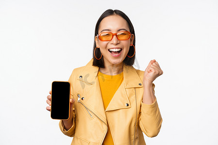 戴墨镜的快乐亚洲女孩，展示手机屏幕、智能手机界面，笑着笑着，庆祝着，站在白色背景上