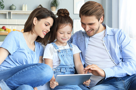 周末在家放松时，积极友好的年轻父母带着微笑的小女儿坐在沙发上，一起在数字平板电脑上接听视频电话。