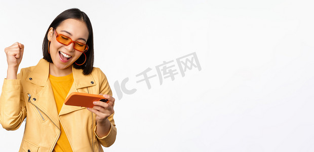 游戏应用摄影照片_戴墨镜的时尚韩国女孩，玩手机视频游戏，在使用智能手机时笑着笑着，站在白色背景上