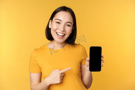 手机小应用摄影照片_美丽快乐的亚洲女孩展示手机屏幕，在智能手机小工具上的应用，站在黄色背景上