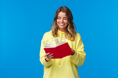 女孩买了新的计划书，写下笔记或演讲，开心地微笑，拿着红色笔记本，决定开始秘密日记，站在蓝色背景的黄色连帽衫