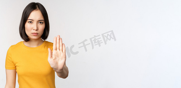 亚洲女性的肖像看起来严肃而愤怒，展示停止禁止手势、禁忌标志、禁止 smth，站在白色背景上的黄色 T 恤