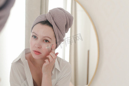 头上戴着毛巾的年轻女子照镜子，在脸上涂抹奶油