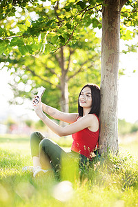 快乐的深棕色头发女孩，她的智能手机在树下
