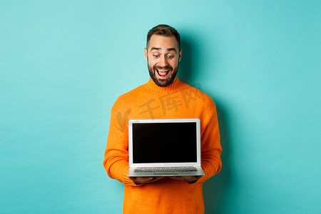 兴奋的成年男性模特展示笔记本电脑屏幕，展示在线促销，在浅蓝色背景下开心地站着