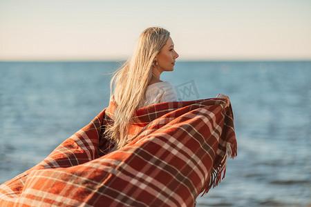 迷人的金发白种女人在日落时分在海滩上享受时光，裹着毯子走着，看着旁边，背景是落日的天空和大海。