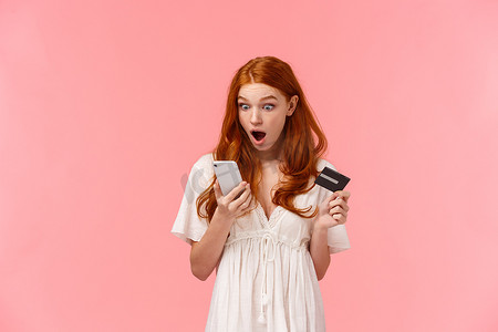 震惊和吃惊的年轻白人红发女孩通过智能手机应用程序看到她银行账户上的东西摇晃，拿着手机和信用卡，低着下巴盯着显示屏