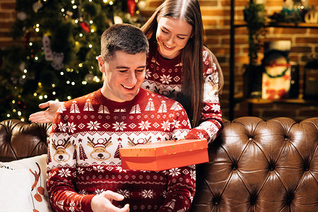 寒假摄影照片_30 多岁的年轻女性遮住微笑好奇的丈夫的眼睛，在装饰过的节日树附近送上装有圣诞礼物的包装盒，新年寒假家庭庆祝活动。