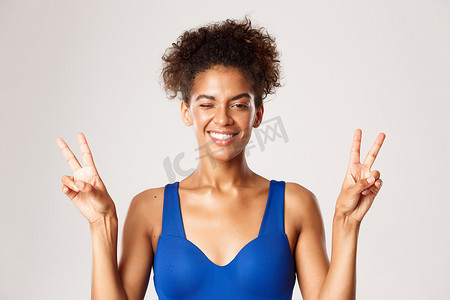 穿着蓝色运动服的快乐的非洲裔美国健身女孩，站在白色背景上，展示和平标志，微笑着开心