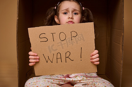 身穿家居服的白人儿童坐在纸箱内，纸箱内贴有“停止战争”海报。