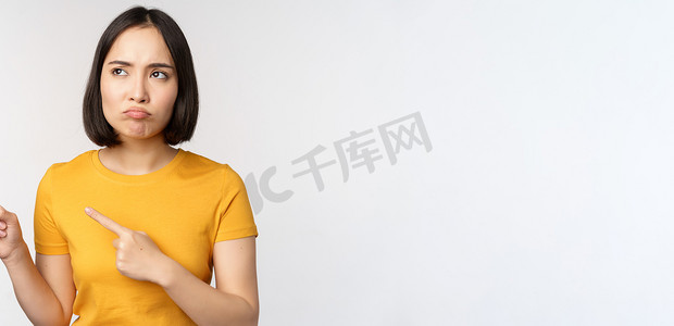 标志品牌摄影照片_脾气暴躁的亚洲女孩心烦意乱，噘嘴，看着不公平的东西，用手指指着横幅、标志品牌，站在白色背景的黄色 T 恤上