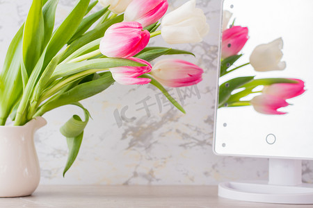 一束带绿叶的白色和粉色郁金香放在桌子上的一个罐子里，映在镜子里。