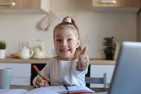 抽签摄影照片_快乐的小女孩学生在家里使用笔记本电脑在线学习，微笑的小孩竖起大拇指推荐课程或课程。