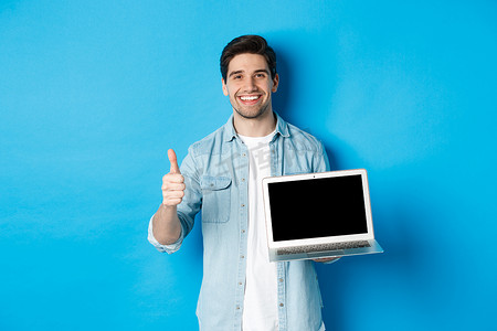微笑的帅哥展示笔记本电脑屏幕和竖起大拇指，比如促销优惠，推荐网站，站在蓝色背景下