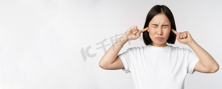亚洲女性的画像闭上耳朵，因噪音、烦人的声音、站在白色背景上而感到不适