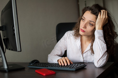 一头卷发和白衬衫的疲惫的年轻女性正在办公室使用她的笔记本电脑、日常工作、自由职业者、倦怠综合症