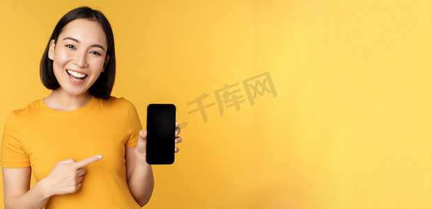 手机小应用摄影照片_美丽快乐的亚洲女孩展示手机屏幕，在智能手机小工具上的应用，站在黄色背景上