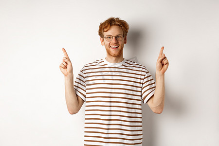 戴着书呆子眼镜的风趣的年轻红头发人手指横着指着，展示两个促销优惠，在白色背景中快乐地站着