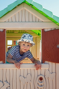 窗外操场摄影照片_一个戴着帽子的快乐小男孩，在一个玩具小屋里，他看着窗外，百叶窗直接对着镜头。