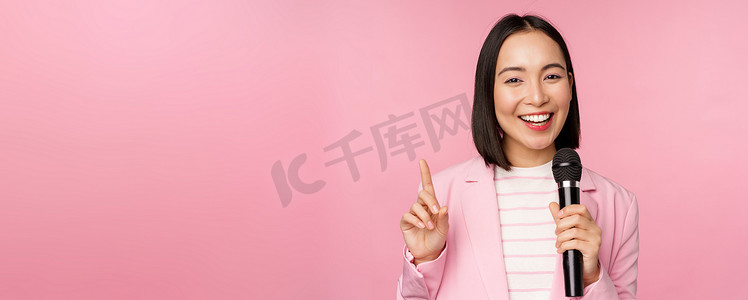 热情的亚洲女商人发表演讲、与麦克风交谈、手持麦克风、穿着西装站在粉红色工作室背景下的形象
