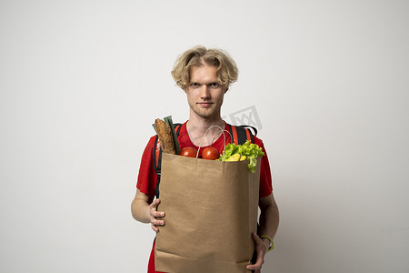 外卖产品背景摄影照片_身穿红色 T 恤制服的送货员拿着牛皮纸包，食物在白色背景工作室中被隔离。