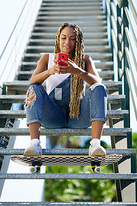 扎辫子女孩摄影照片_扎着彩色辫子的黑人女性，双脚搁在滑板上查看她的智能手机。