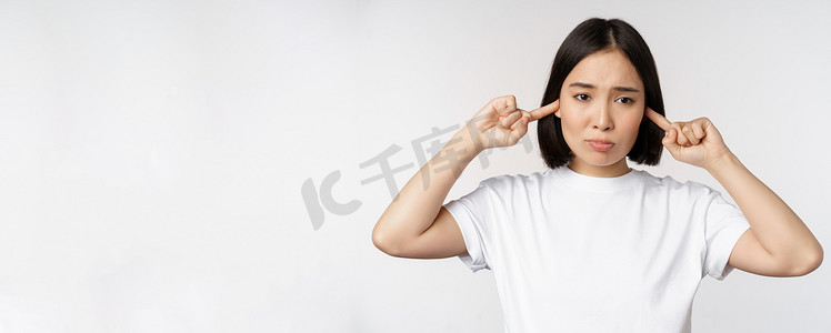 烦人摄影照片_亚洲女性闭上耳朵，站在白色背景上，因巨响、烦人的声音而感到不适的肖像