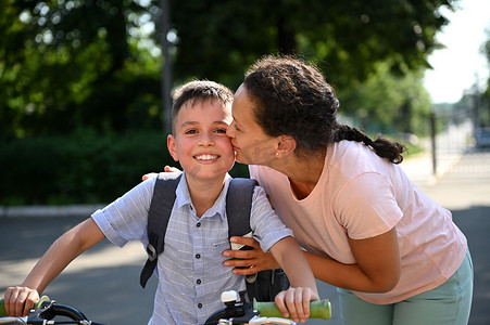 男孩骑自行车上学摄影照片_快乐的母亲拥抱并亲吻她儿子的脸颊，带他去上学。