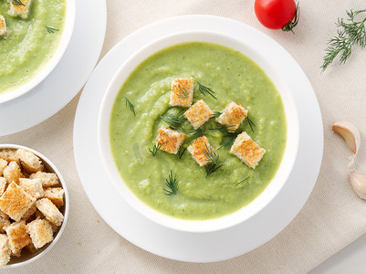 两个大白碗，上面有西兰花、西葫芦、绿豌豆的蔬菜绿色奶油汤，白色背景，顶视图
