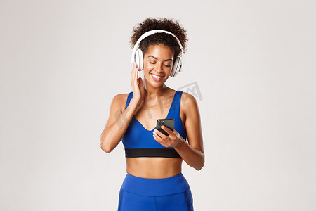 穿着蓝色运动服的漂亮健身女性，用无线耳机听音乐锻炼，使用智能手机应用程序，站在白色背景下