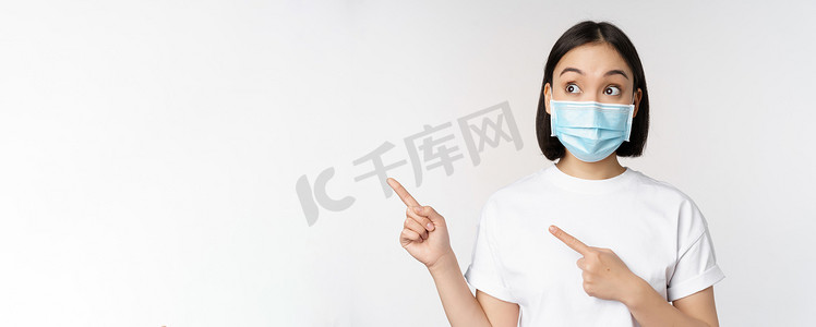 戴着医用面罩的年轻韩国女性手指向左，看着标志，展示广告或横幅，站在白色背景上