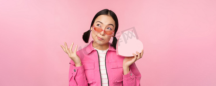 粉色心形礼物盒摄影照片_戴着太阳镜的时尚亚洲女友的形象，猜猜礼盒里有什么，心形礼物，站在粉色背景上