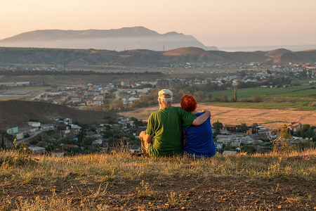 一对老夫妇背坐在山上，眺望着远处的山海美景。