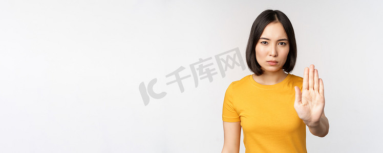 亚洲女性的肖像看起来严肃而愤怒，展示停止禁止手势、禁忌标志、禁止某物、站在白色背景上的黄色 T 恤