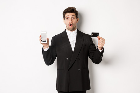 穿着黑色西装的英俊商人的形象，看起来很惊讶，并用手机屏幕展示信用卡，站在白色背景下