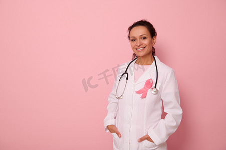 国际卫生组织摄影照片_非洲裔美国医生的画像，身穿带粉红丝带的医用大衣，脖子上挂着听诊器，看着相机，在彩色背景中被隔离，复制空间。