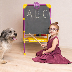 学生给老师摄影照片_一位穿着裙子、戴着眼镜的小女老师坐在黑板附近的地板上，给她的狗看英文字母。